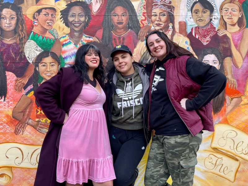 Three women posing in front of a mural of women at Balmy Alley and Lovers Lane.