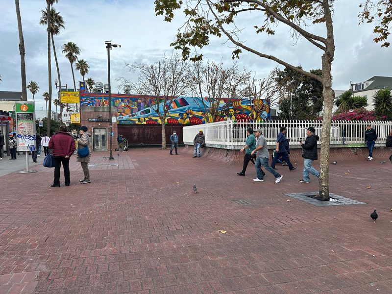 A group of people walking on a brick sidewalk at 24th Street BART Plaza