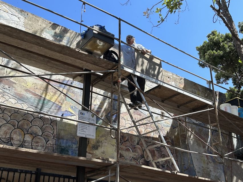 A man scales a two-story scaffolding in front of a mural he is restoring.