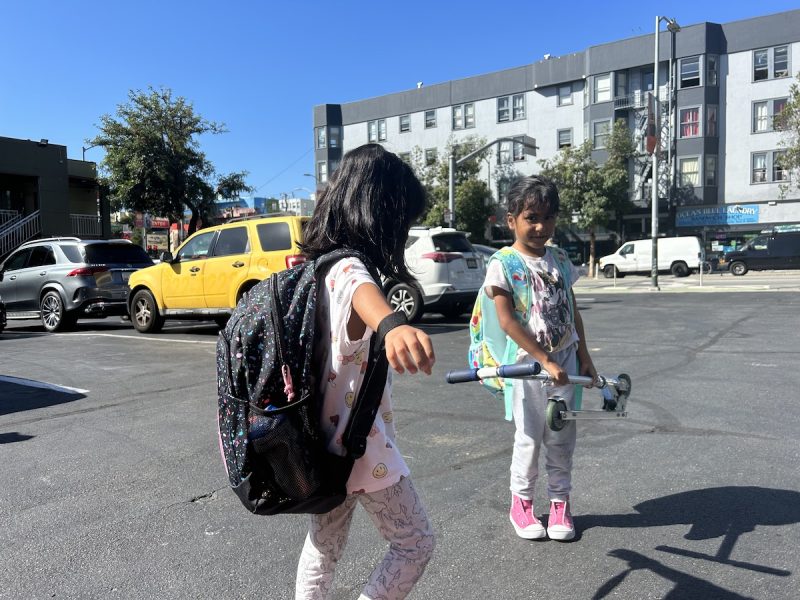 Two young girls play on a blacktop and have backpacks on. One is holding a folded-up Razor scooter.