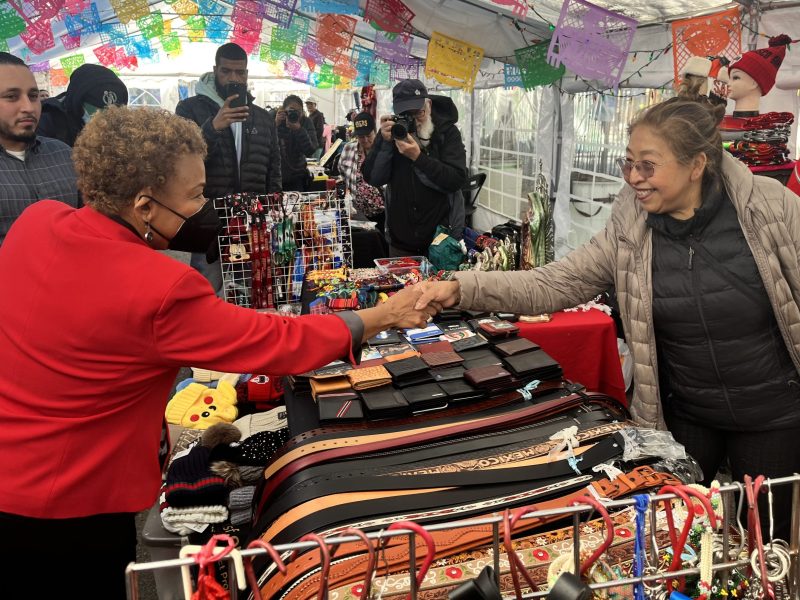Rep. Barbara Lee shaking hands with a vendor at La Placita, the new space for street vendors at 24th and Capp, in front of a table filled with belts, wallets, and other goods.