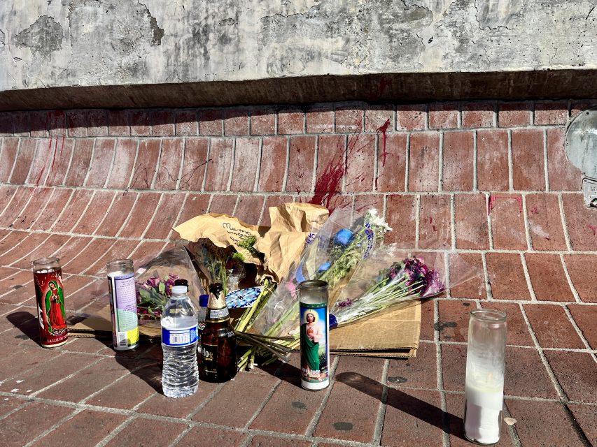 A group of candles, flowers, and a bottle of water on a brick wall in a memorial for Guillermo.