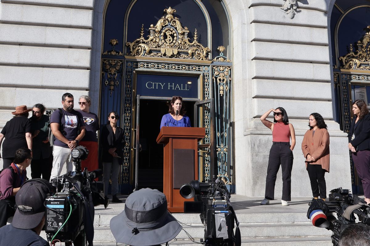 A group of people gather around a podium in front of City Hall.