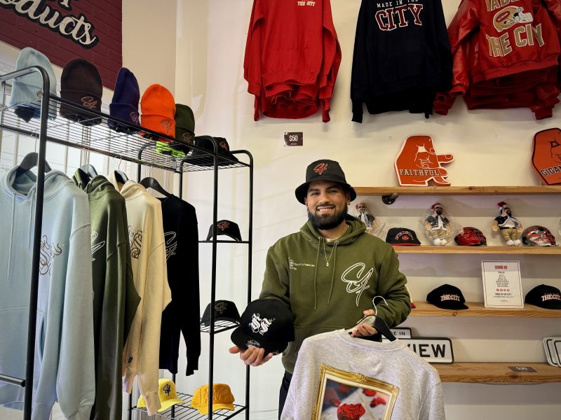 A man standing in front of a rack of hats, sweatshirts and hoodies.