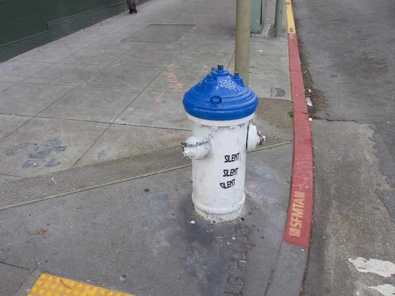 A blue and white fire hydrant on a sidewalk. The word silent is stenciled on to it three times.