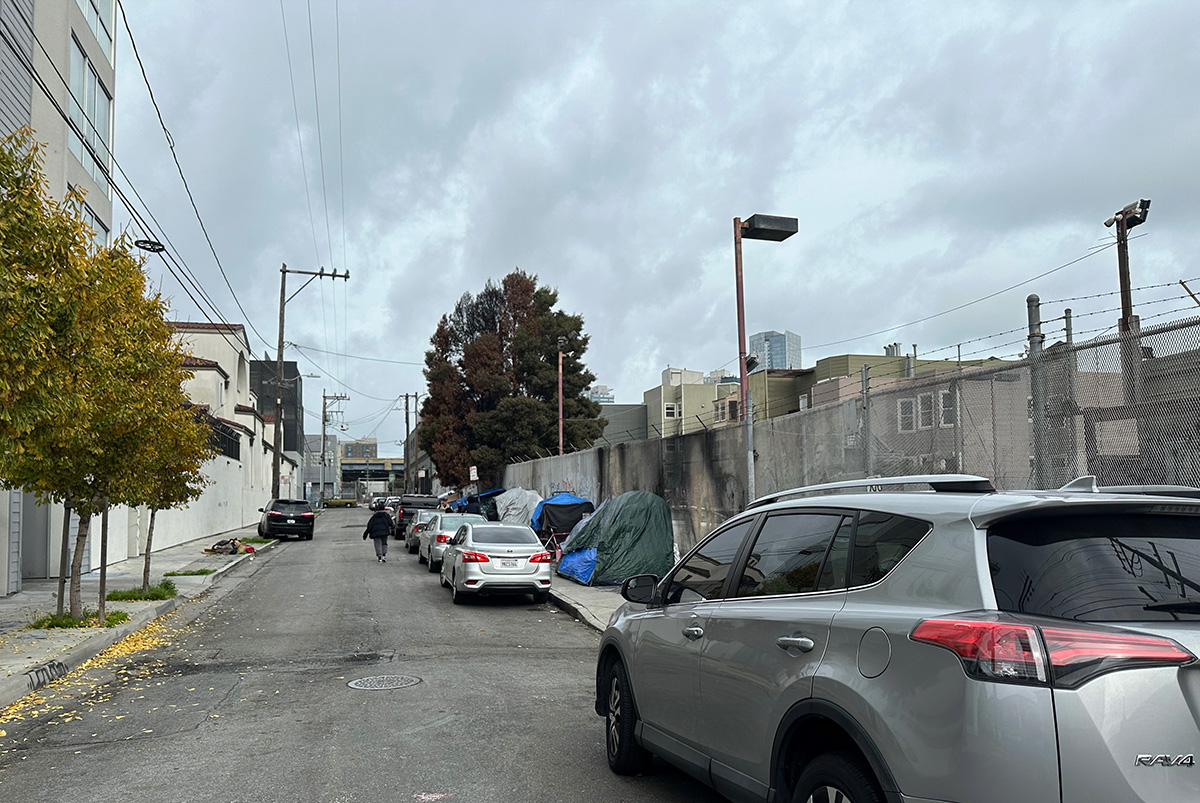 A street with cars parked next to a fence, adjacent to an encampment.