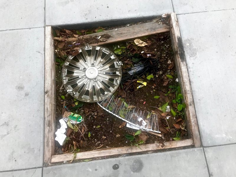 A wheel is sitting in a planter on the sidewalk.