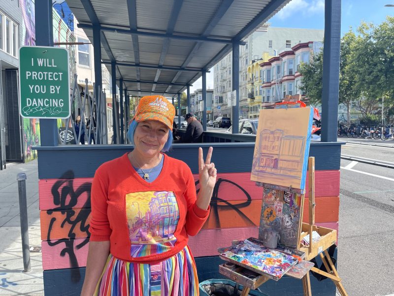 Jirsa, rainbow skirt billowing in the wind, stands in their rainbow corner, painting the buildings across the street.