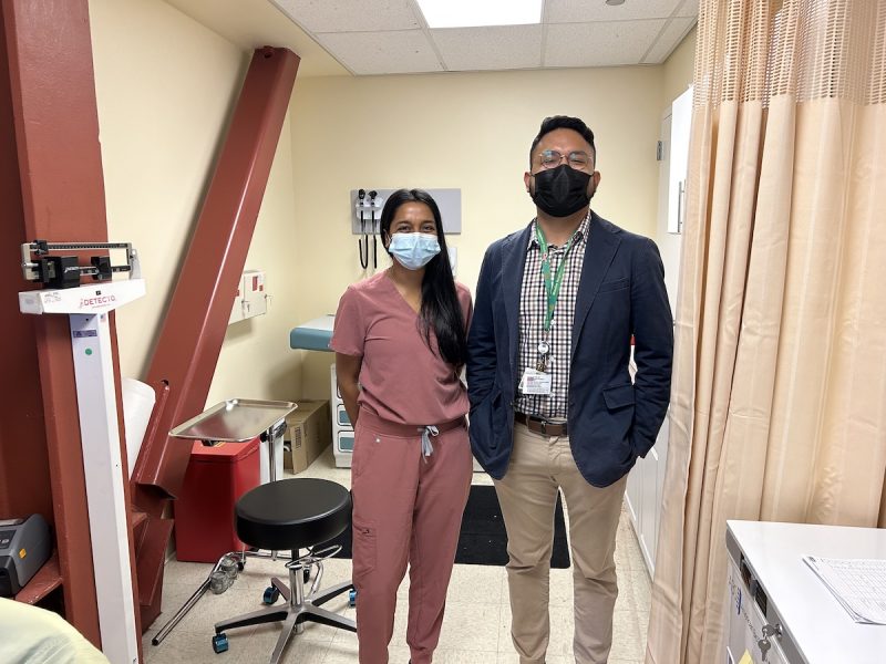 A pediatrician dressed in blush scrubs and a mask, and a health center representative in a blue blazer, pose in front of a brand-new clinic at a high school.
