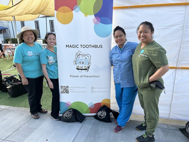 Four women pose in front of a 'Magic ToothBus' sign. Two are in scrubs.