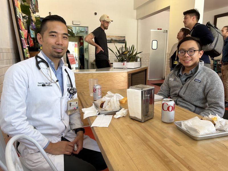 At Newkirk's cheesesteak sandwich shop, a hospital doctor dressed in a white coat and stethoscope and a resident eat at a wooden table.