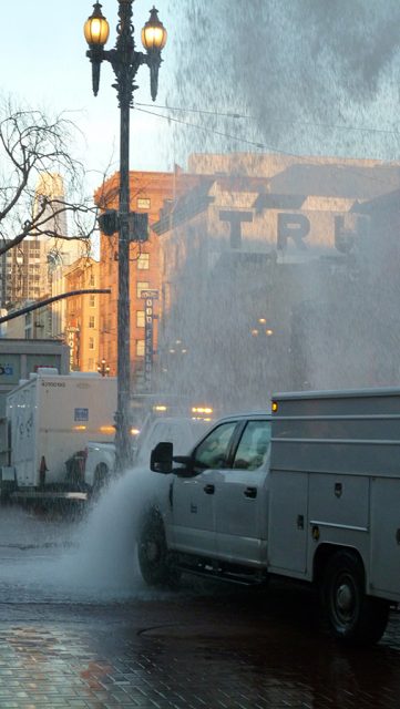 A truck driving down a street with a water hose coming out of it.
