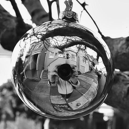 The reflection of.a man's hat an body in a Christmas bulb hanging from a tree outside.