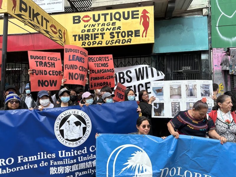 Multiple people of different races stand under a yellow awning and brandish colorful posters that demand funding for tenant programs.