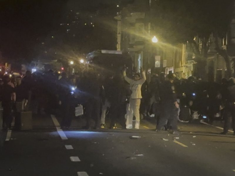 A night shot of teenagers with hands up, officers, vans after the 2023 Dolores Park hill bomb