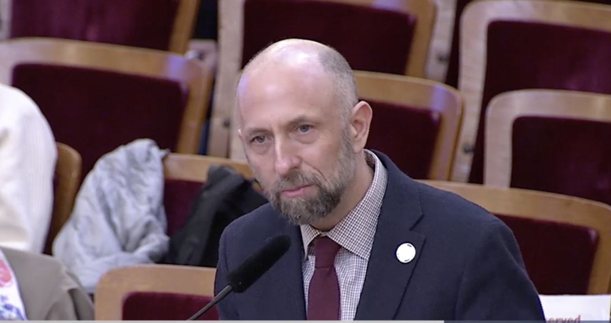 A bald man in a suit speaking into a microphone.